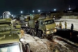 Green military vehicles drive off a railway platform and on to snowy roads while soliders walk about.