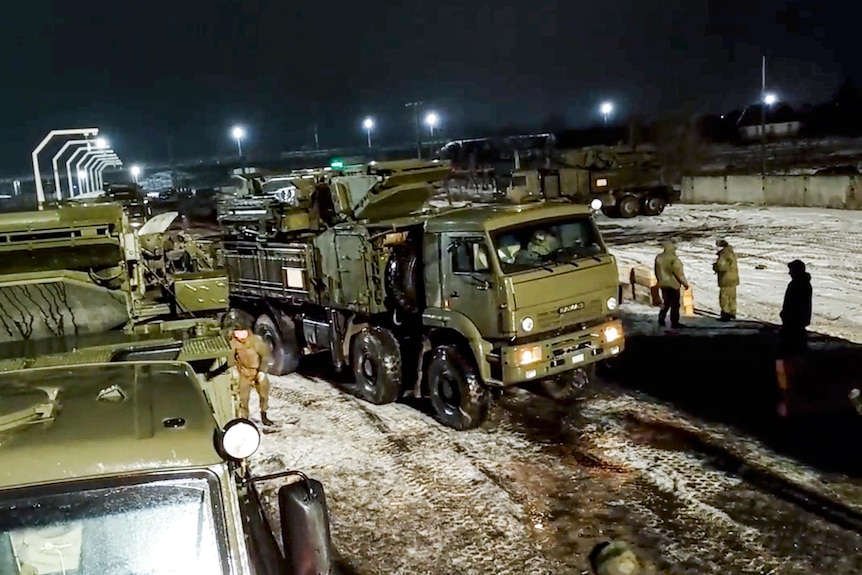 Green military vehicles drive off a railway platform and on to snowy roads while soliders walk about.