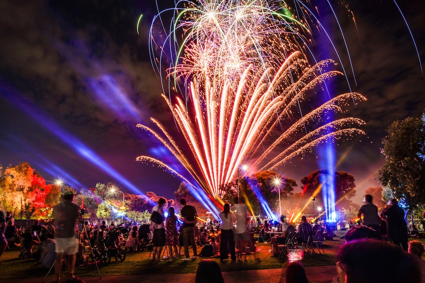Bright orange fireworks and blue and purple laser beams cascade out over a park with people watching at night 
