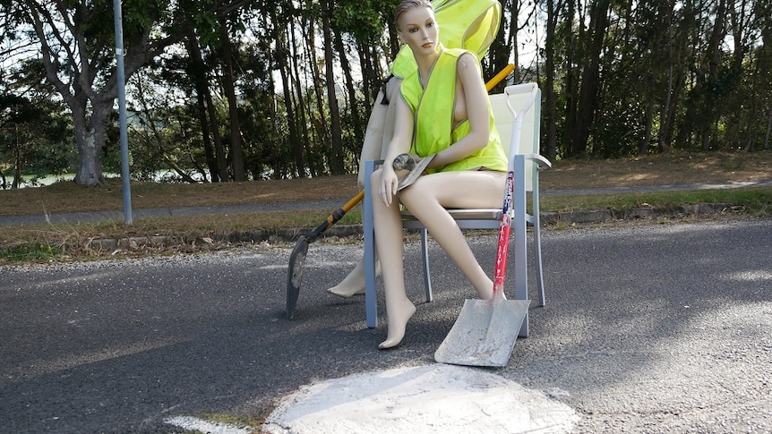 Two mannequins in high vis vests and shovels stand and sit by a pothole which has been illegally filled