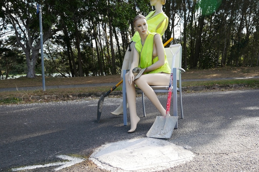 Two mannequins in high vis vests and shovels stand and sit by a pothole which has been illegally filled