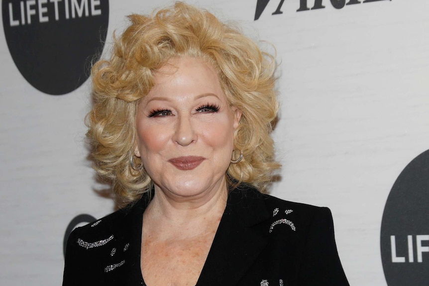 Bette Midler smiles for the camera wearing a black suit with sequin smiley faces on the jacket.