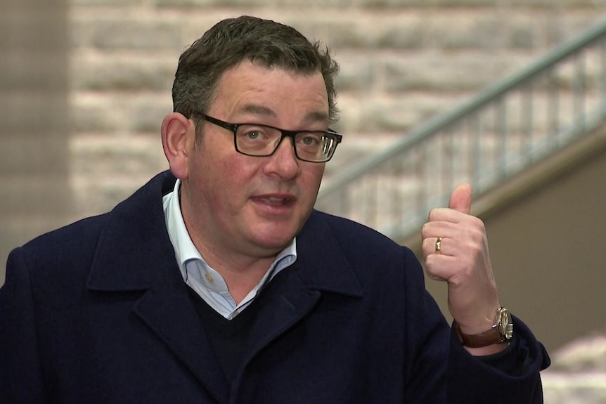 Daniel Andrews, dressed in a shirt and coat, gestures with his hand as he speaks at a press conference.
