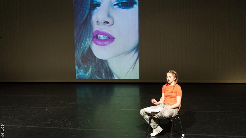 Samira Elagoz sitting on a chair on a stage wearing an orange shirt