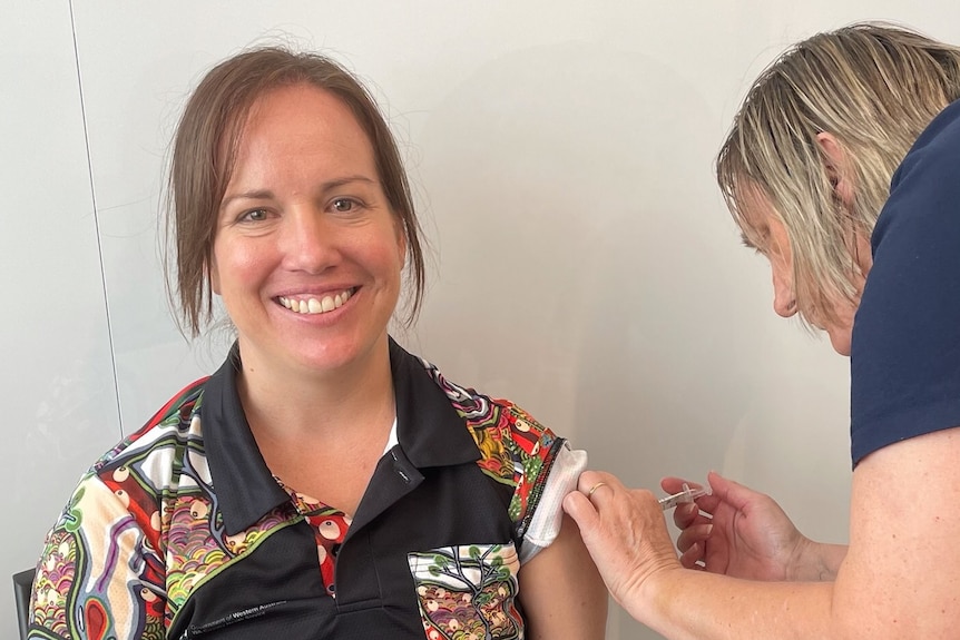 A woman smiles at the camera as she receives an injection in her arm