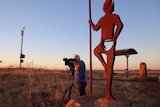 Cameraman Robert Koenig-Luck standing next to giant indigenous statues while filming the landscape of Roebourne at sunset.