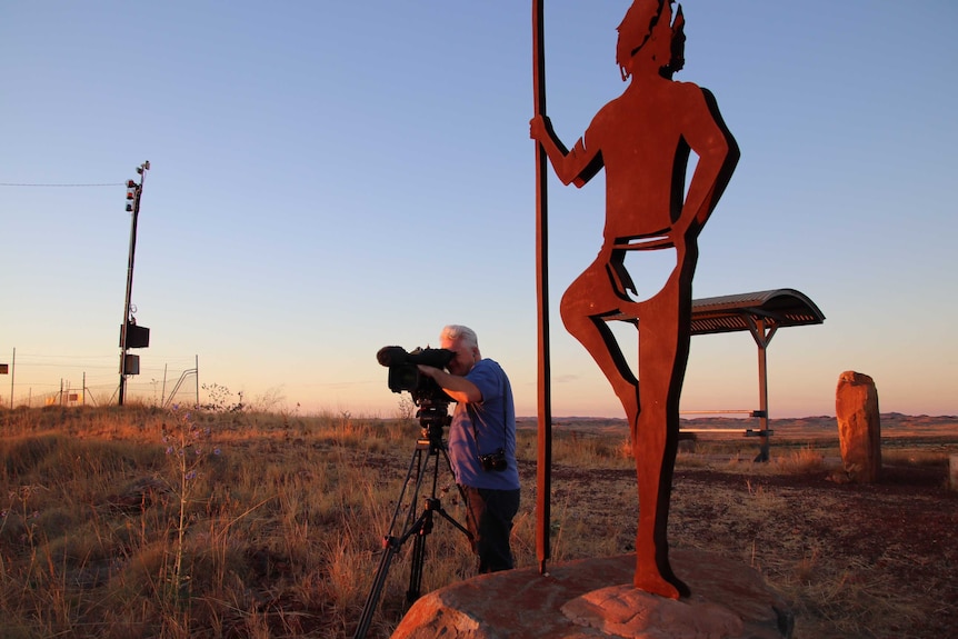 Cameraman Robert Koenig-Luck standing next to giant indigenous statues while filming the landscape of Roebourne at sunset.