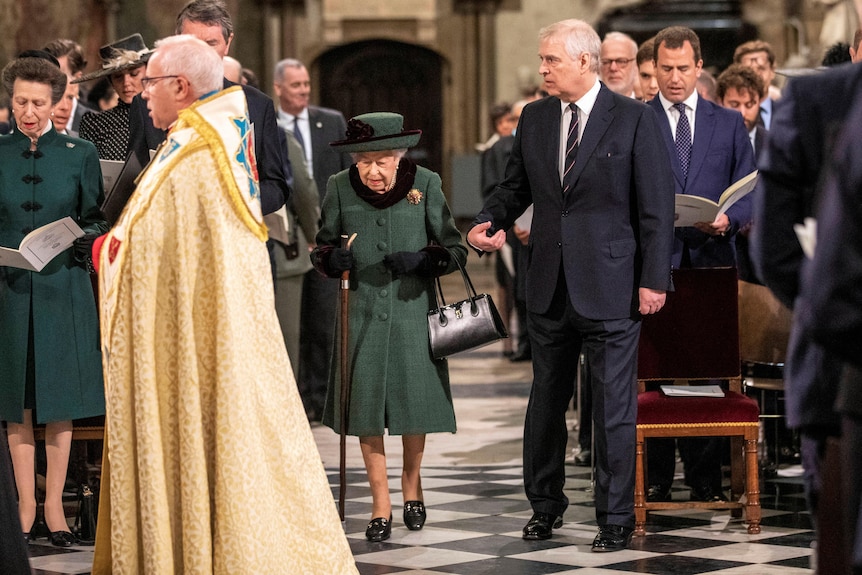 Prince Andrew in a suit offers his arm to his mother dressed in green as they walk down an aisle.