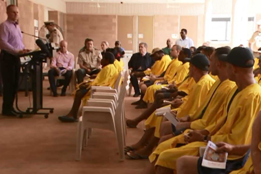 Prisoners sit in yellow uniforms as man talks at a lectern