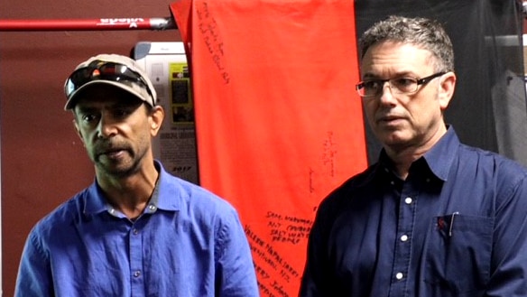 Two Aboriginal men stand in front of an aboriginal flag with writing on it.