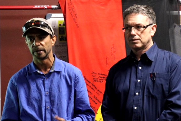 Two Aboriginal men stand in front of an aboriginal flag with writing on it.