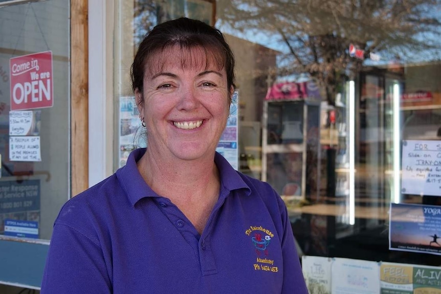 A smiling woman with brown hair wearing a purple polo shirt and standing outside a bakery.