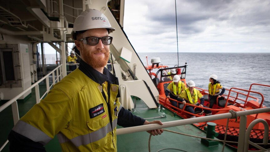 Man in glasses and helmet on deck of vessel at sea, life raft on ship in background with crew on it
