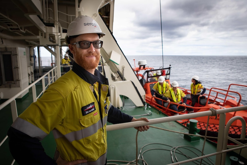 Man in glasses and helmet on deck of vessel at sea, life raft on ship in background with crew on it