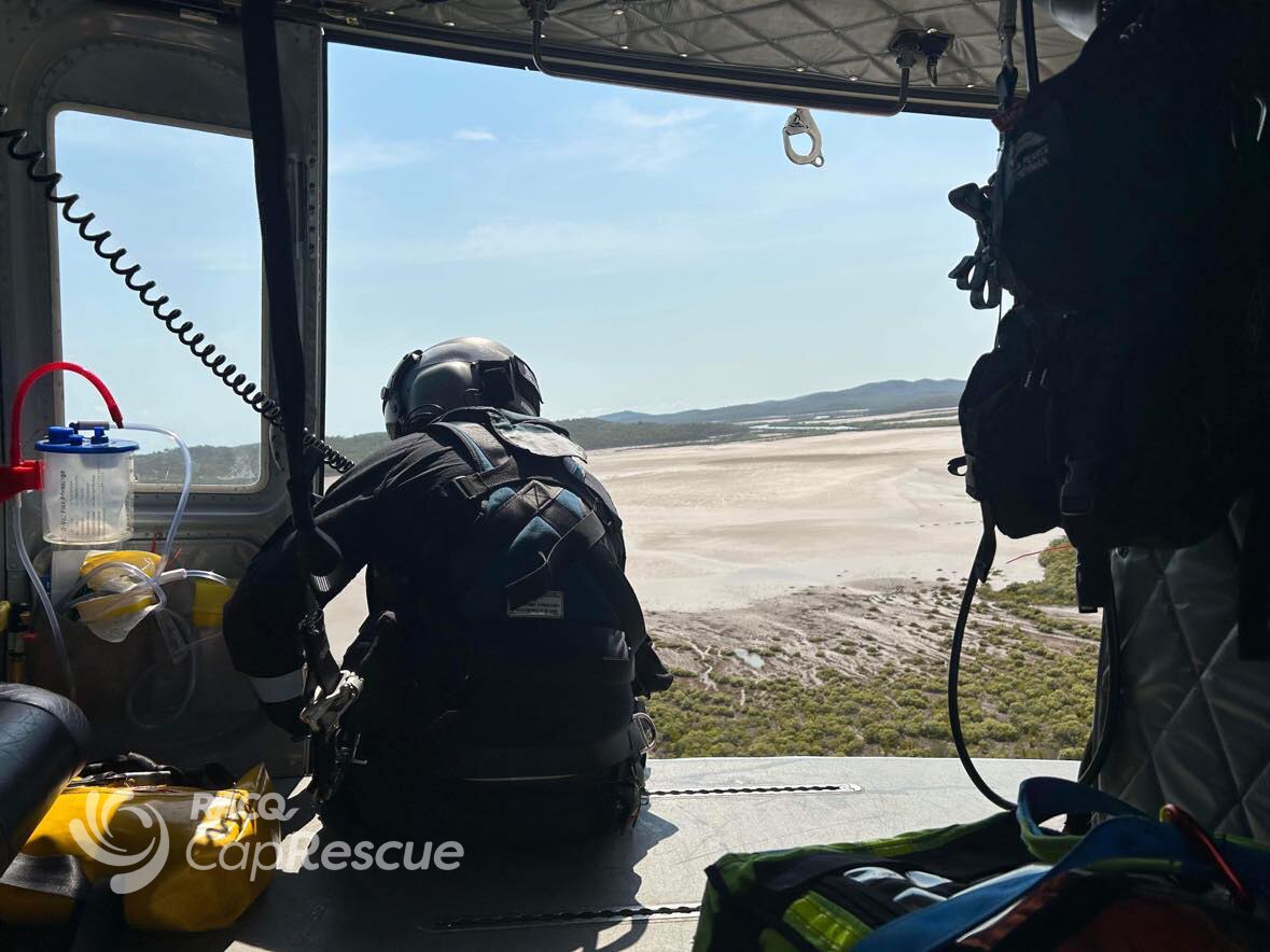 Man wearing protective gear leans out the side of a helicopter in the air over a beach.