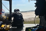 Man wearing protective gear leans out the side of a helicopter in the air over a beach.