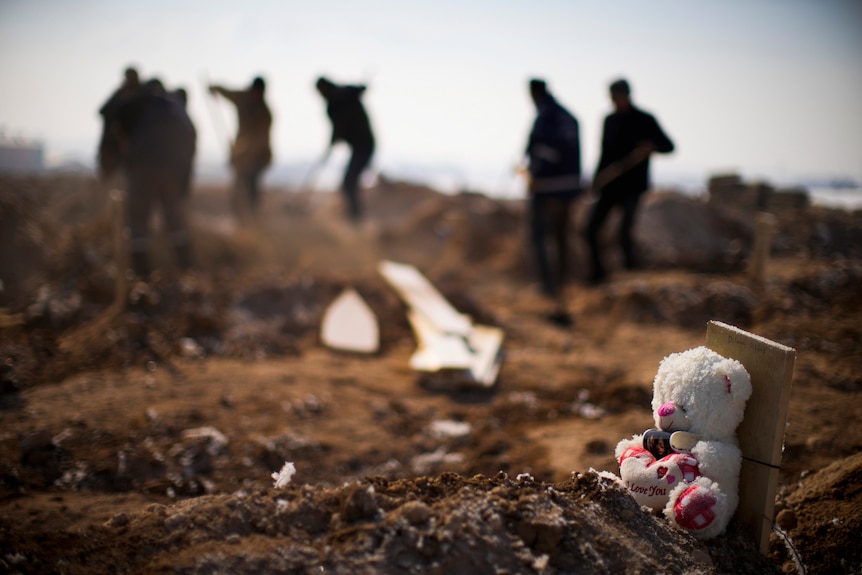 A teddy bear sits on a grave as people dig more graves in the background.