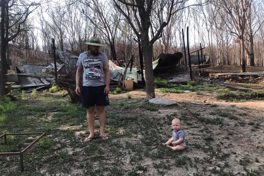 A man stands in front of a burnt out house with his toddler on the ground