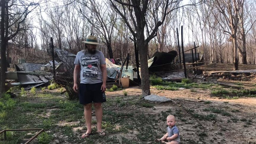 A man stands in front of a burnt out house with his toddler on the ground