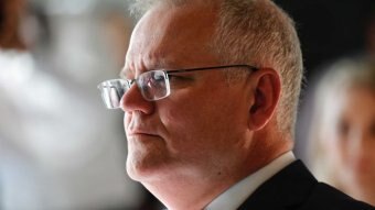 Prime Minister Scott Morrison looks to the left in portrait image. A reflection is visible in his glasses and he wears a suit.