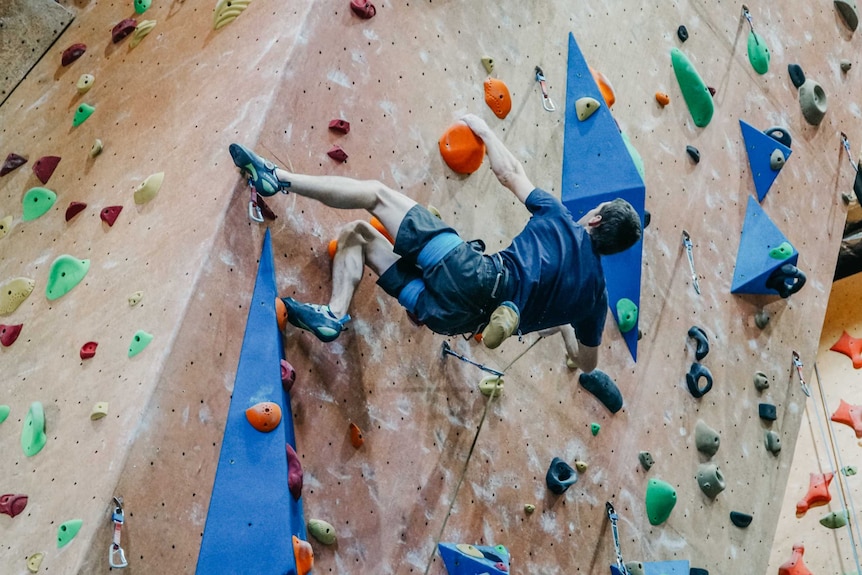 A man hangs by one arm from a colourful indoor rock climbing wall.