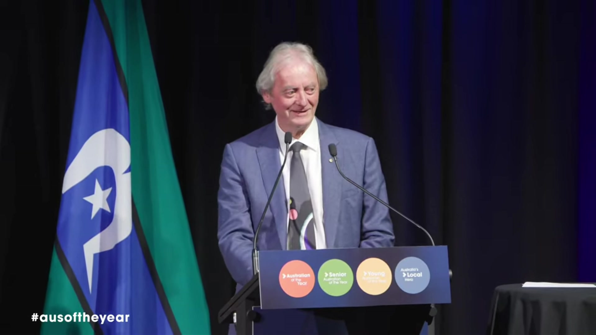 A man with sandy hair in a blue jacket, white collared shirt and black tie stand on stage behind microphones.