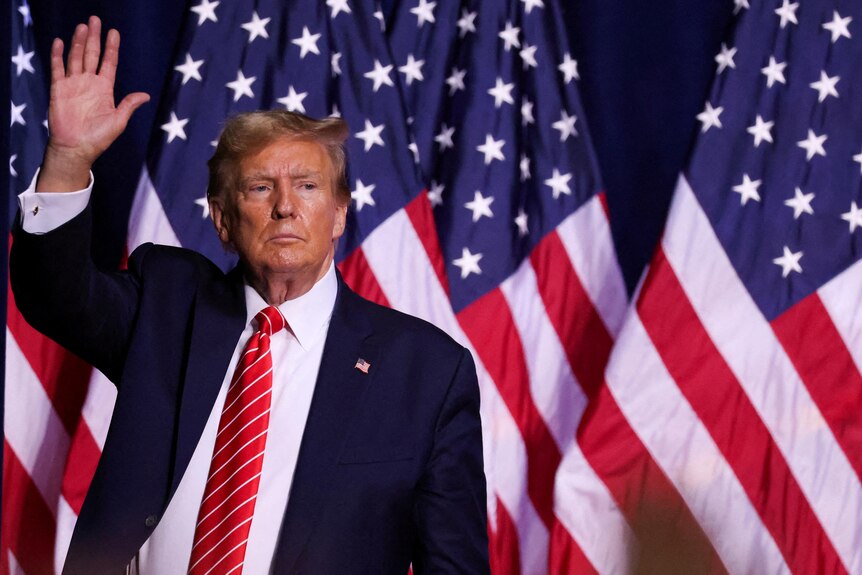 Donald Trump wearing a suit with a red tie raises his hand while standing in front of a United States flag.