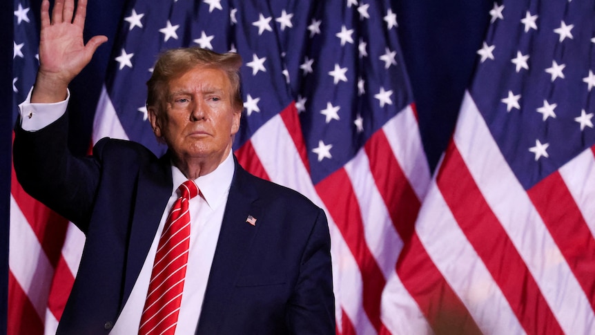 Donald Trump wearing a suit with a red tie raises his hand while standing in front of a United States flag.