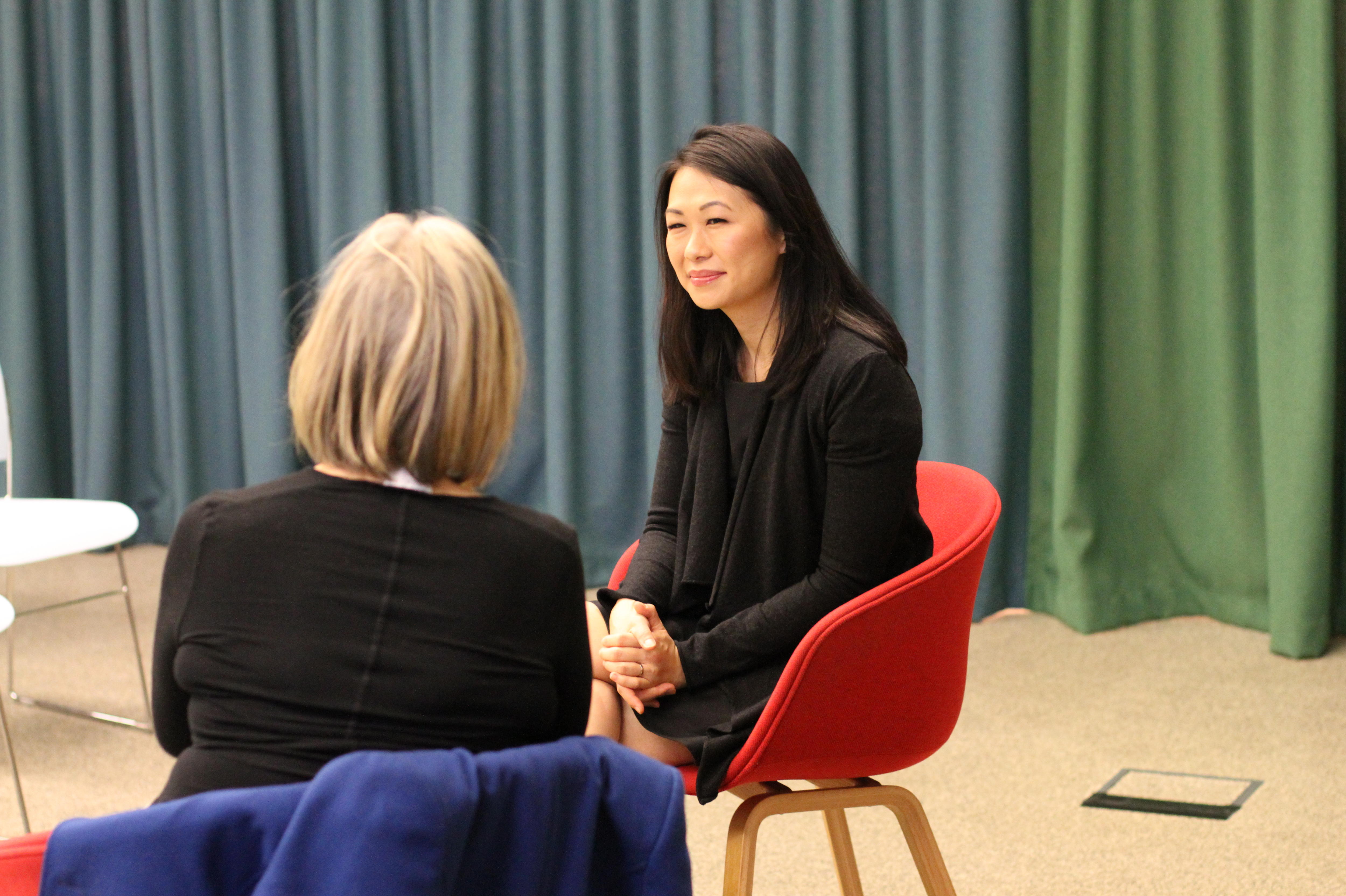 A woman with long dark hair sits smiling and talking to another woman.