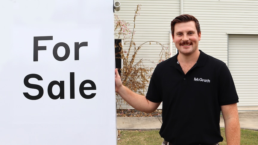 Man smiles next to a for sale sign