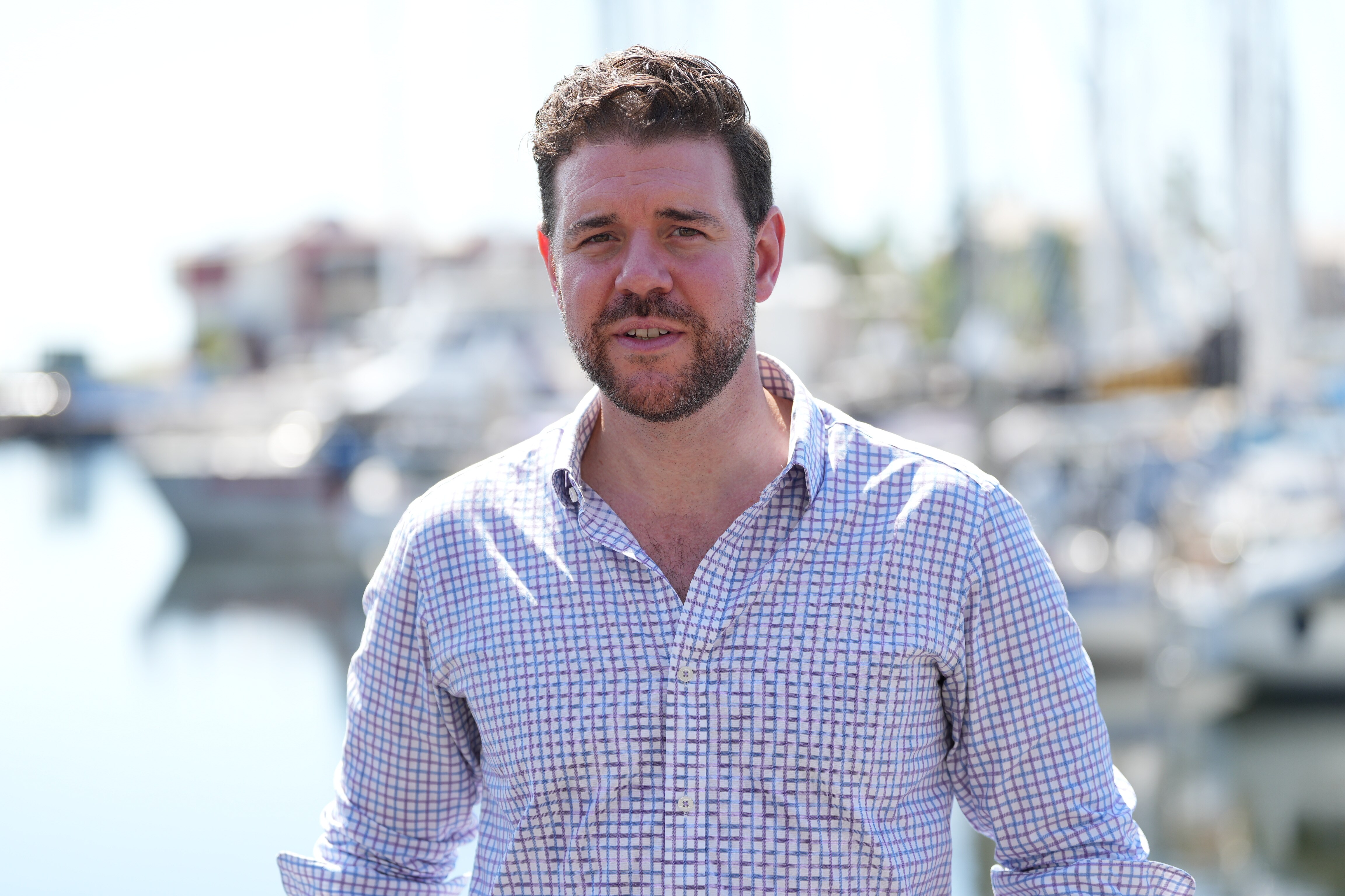 Un hombre blanco, castaño, espeso, pelo corto, barba, vestido con una camisa de cuadros azules y blancos, el puerto se difumina detrás de él.