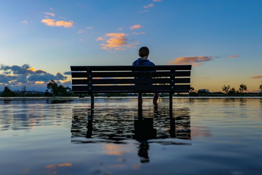A photo of a child sitting on a bench in front of a river with a puddle underneath reflecting the sky.