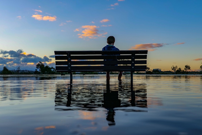 A photo of a child sitting on a bench in front of a river with a puddle underneath reflecting the sky.