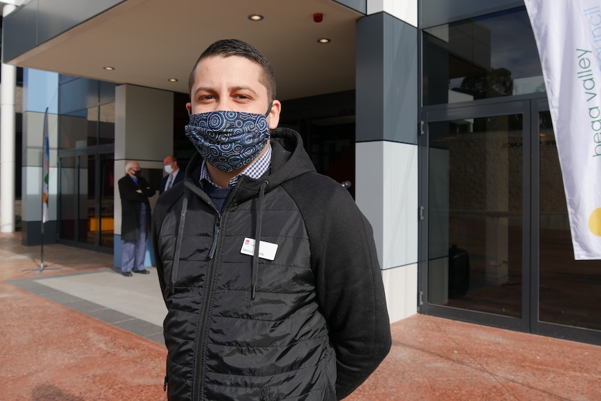 A man in a dark jacket stands with an Indigenous designed mask on his face in front of a building