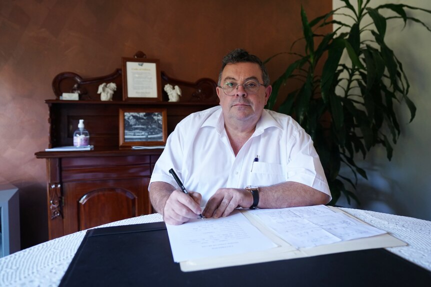 A man sits in his office wearing a white shirt and glasses looking a paperwork