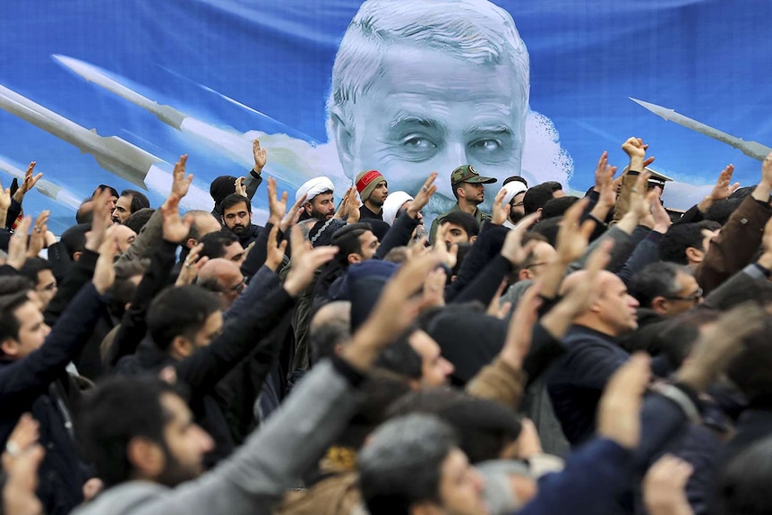 A crowd of people stand with one hand raised about their heads. Behind them is a banner displaying General Soleimani's face