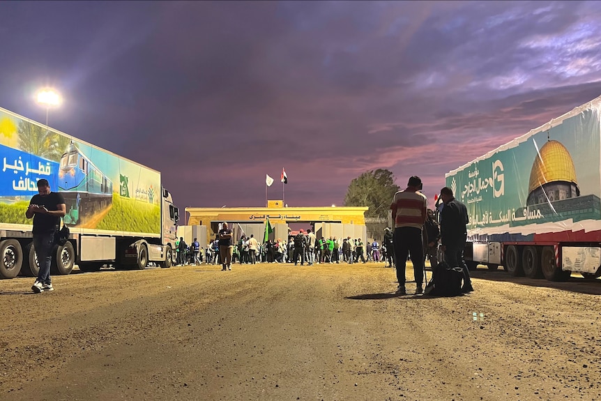 Two trucks are parked in front of a checkpoint-type building, as people walk past and the sun sets