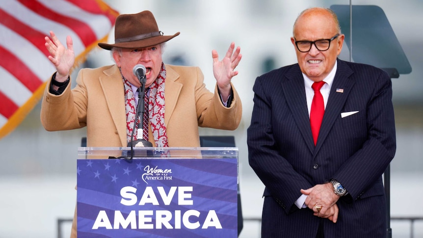 John Eastment stands behind a podium gensturing with his hands wearing a hat. Rudy Giuliani is next to him smiling