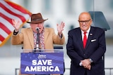 John Eastment stands behind a podium gensturing with his hands wearing a hat. Rudy Giuliani is next to him smiling