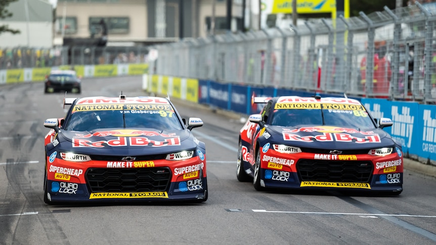 Two Red Bull Chevrolet Supercars race side by side down a straight with a metal fence and barricade to the right.
