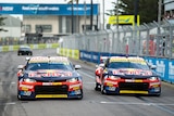 Two Red Bull Chevrolet Supercars race side by side down a straight with a metal fence and barricade to the right.