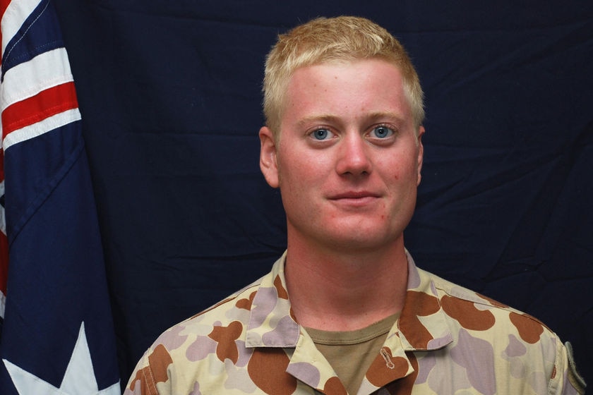 A blond-haired young man in his army fatigues stands in front of an Australian flag