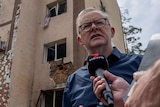 Albanese speaks into a microphone being held by a journalist, with destroyed buildings behind him.