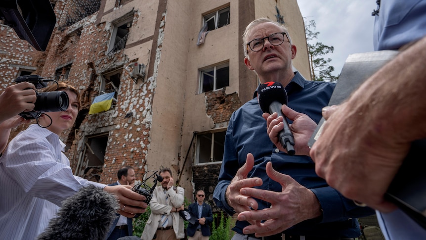 Albanese speaks into a microphone being held by a journalist, with destroyed buildings behind him.