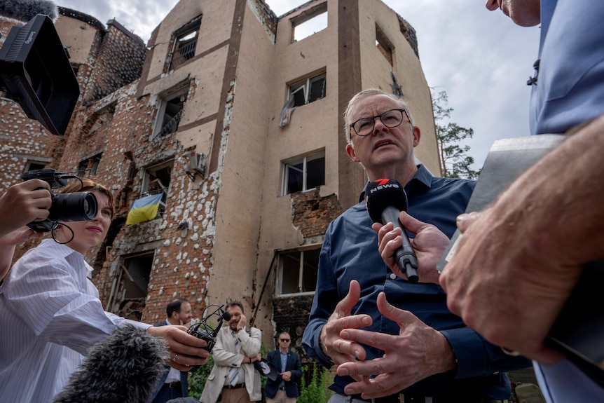 Albanese speaks into a microphone being held by a journalist, with destroyed buildings behind him.