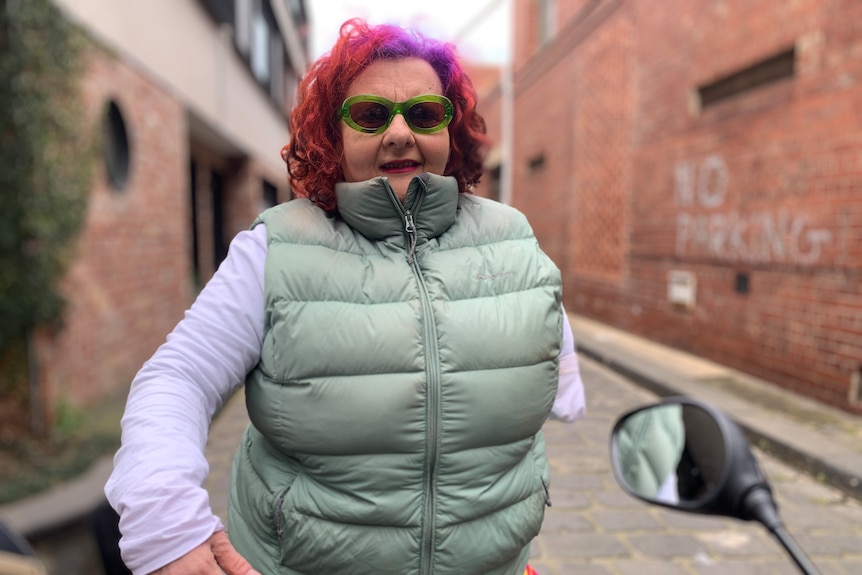 An older woman with red hair standing in a laneway. She is wearing a grey puffer vest