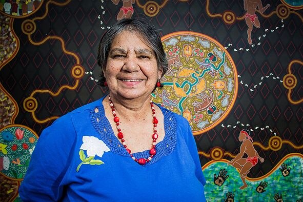 A smiling short-haired older woman in front of indigenous painting, blue top with flower on right, red necklace.