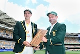 Pat Cummins and Dean Elgar stand in green blazers holding a wooden trophy