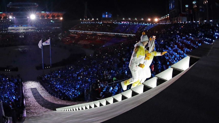Wide shot of two people carrying a torch up stairs with a large crowd behind them.