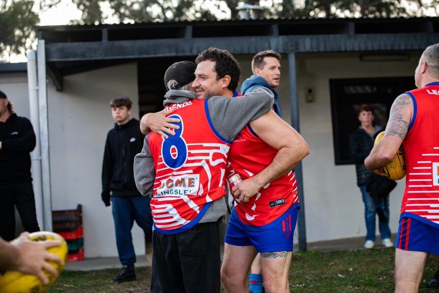A wide shot of Brad hugging another man whose back is facing the camera 